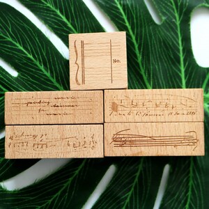 Puede incluir: Cinco sellos de madera con diferentes diseños. El sello superior tiene una nota musical y el texto "No.". El segundo sello tiene el texto "painting music for whatever music". El tercer sello tiene un patrón de cuadrícula con el texto "1. Sonata (C.P.E. Bach) H.588". El cuarto sello tiene un diseño de nota musical. El quinto sello tiene un diseño de nota musical.