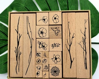 May include: A set of 20 wooden stamps featuring floral and branch designs. The stamps are arranged in a wooden box with a light brown background. The stamps are all different sizes and shapes, and they are all made of natural wood.