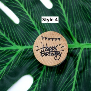 Puede incluir: Un sello de madera con el texto "Happy Birthday" escrito a mano. El sello tambi&eacute;n presenta una hilera de banderas blancas y negras.