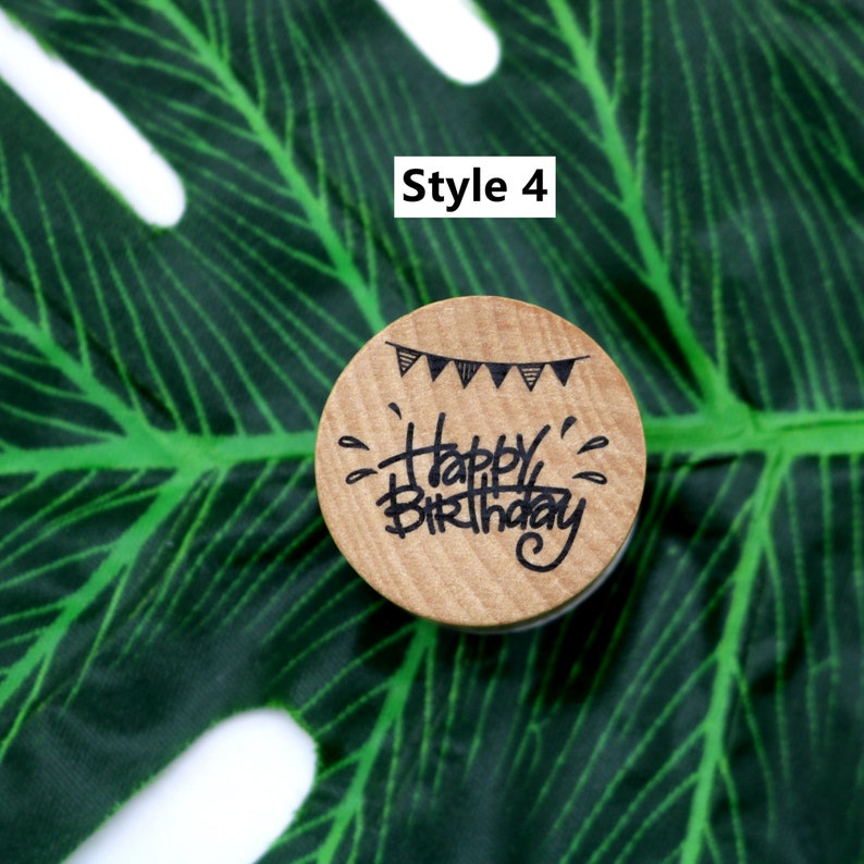 Puede incluir: Un sello de madera con el texto "Happy Birthday" escrito a mano. El sello tambi&eacute;n presenta una hilera de banderas blancas y negras.