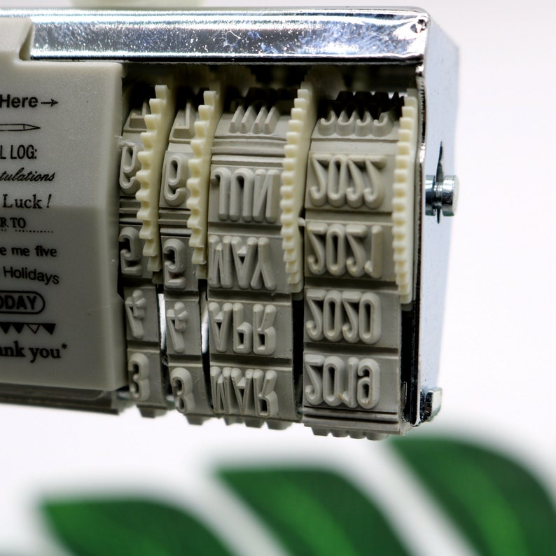 May include: A silver and white date stamp with a series of rotating wheels. The wheels are labeled with the numbers 1, 2, 3, 4, 5, 6, 7, 8, 9, 0, and the letters SOS, YAN, and TIME.