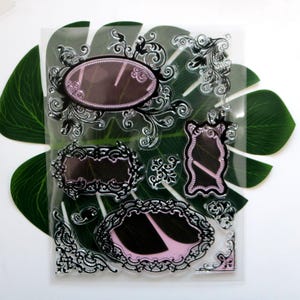 May include: A clear stamp set showcasing various ornate black frames with pink and black interiors. The frames are in oval, rectangular, and arched shapes. The set includes decorative flourishes and corner accents, suitable for scrapbooking and card making projects.