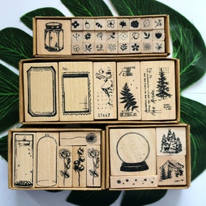 Puede incluir: Sellos de madera en cajas de cartón. Los sellos presentan varios diseños, incluyendo patrones florales, marcos, un tarro, una bola de nieve, un helecho y un paisaje de montaña. Ideales para scrapbooking y manualidades.