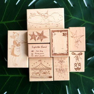 May include: A collection of eight rectangular wooden rubber stamps featuring engraved designs including a child with a tray, a carousel, a chandelier, ballet slippers, stars, a postage frame, a cherub on a cloud, and text reading Inspiration Board.