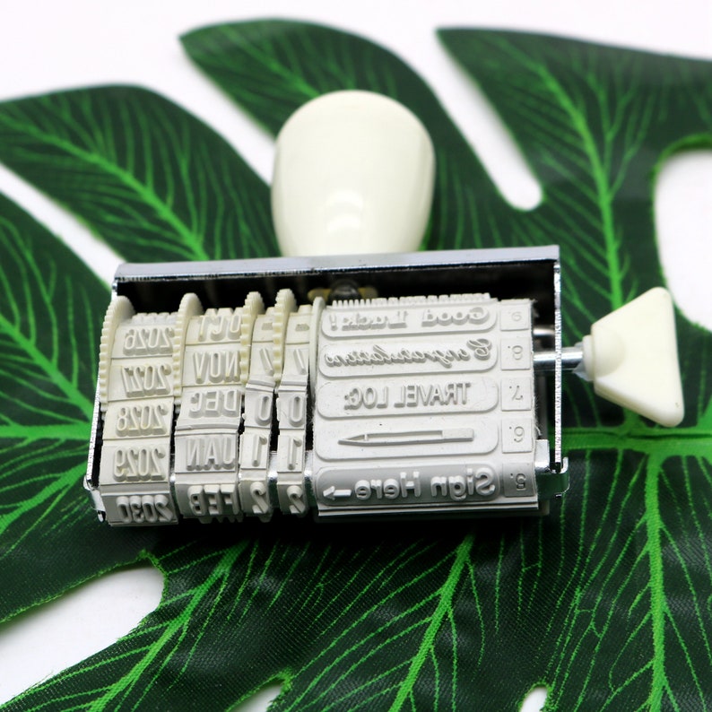 May include: A silver and white date stamp with a handle. The stamp has a series of wheels with numbers and months, and a row of text that reads "Sign Here - Travel Log - Congratulations - Good Luck".