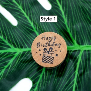 Puede incluir: Un sello de madera redondo con el texto "Happy Birthday" y una peque&ntilde;a caja de regalo con un lazo en la parte superior. El sello est&aacute; sobre un fondo de hoja verde.