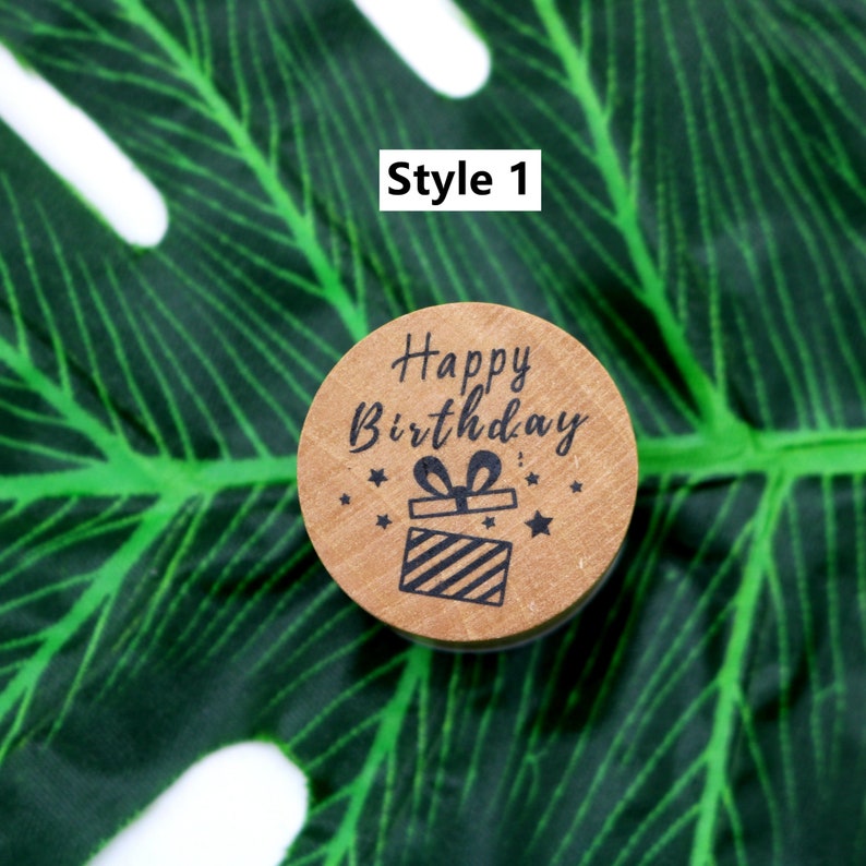 Puede incluir: Un sello de madera redondo con el texto "Happy Birthday" y una peque&ntilde;a caja de regalo con un lazo en la parte superior. El sello est&aacute; sobre un fondo de hoja verde.