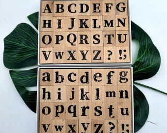 May include: A set of wooden alphabet stamps in a white box. The stamps are arranged in two rows, with uppercase letters in the top row and lowercase letters in the bottom row. The stamps also include punctuation marks like question marks and exclamation points.