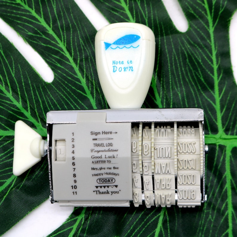 May include: A silver and white vintage rubber stamp with a rotating dial and multiple message options. The stamp has a white plastic top with a blue fish design and the text "Note it Down". The stamp has the following messages: "Sign Here", "Travel Log", "Congratulations", "Good Luck", "A Letter To", "Hey, give me five", "Happy Holidays", "Today", and "Thank you".