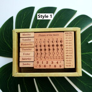 K&ouml;nnte beinhalten: Ein Holzstempel-Set mit Wochentagen, Zahlen und Mondphasen. Die Stempel sind hellbraun mit schwarzer Beschriftung. Die Schachtel ist aus Pappe und hat einen hellgr&uuml;nen Hintergrund.