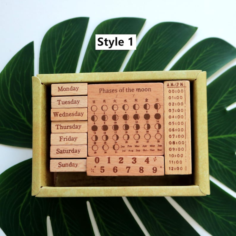 K&ouml;nnte beinhalten: Ein Holzstempel-Set mit Wochentagen, Zahlen und Mondphasen. Die Stempel sind hellbraun mit schwarzer Beschriftung. Die Schachtel ist aus Pappe und hat einen hellgr&uuml;nen Hintergrund.