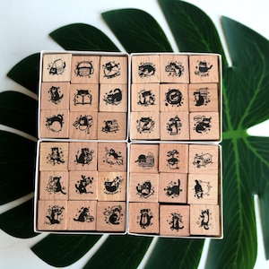 May include: A collection of square wooden stamps arranged in two white boxes. Each stamp features a unique black ink illustration of a cat engaged in various activities, including reading, drinking coffee, and playing with objects, on a light wood base.