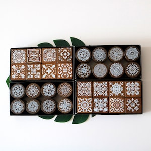 May include: Four black boxes filled with decorative coasters. The coasters feature various white and light blue geometric patterns on a brown background. The boxes are arranged on a white surface with green leaves.