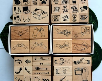 Puede incluir: Un conjunto de sellos de madera con varios diseños, que incluyen gatos, café, comida, manos, flechas y otros símbolos. Los sellos están dispuestos en cuatro filas, cada fila en una caja separada. Las cajas son de madera y tienen un acabado natural.