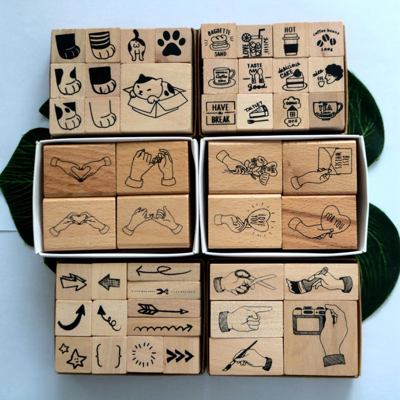 May include: A set of wooden stamps with various designs, including cats, coffee, food, hands, arrows, and other symbols. The stamps are arranged in four rows, each row in a separate box. The boxes are made of wood and have a natural finish.