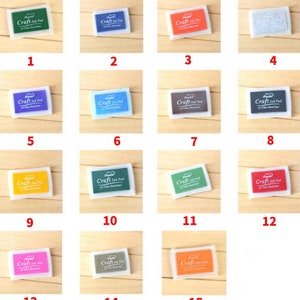 May include: Fifteen small, square, white plastic ink pads with different colored ink inside. The ink pads are labeled "Craft Ink Pad" and are arranged in a grid of five rows and three columns.