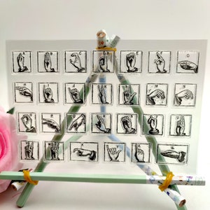 May include: A clear sheet with 26 alphabet letters in American Sign Language. Each letter is in a square frame and is printed in black ink.