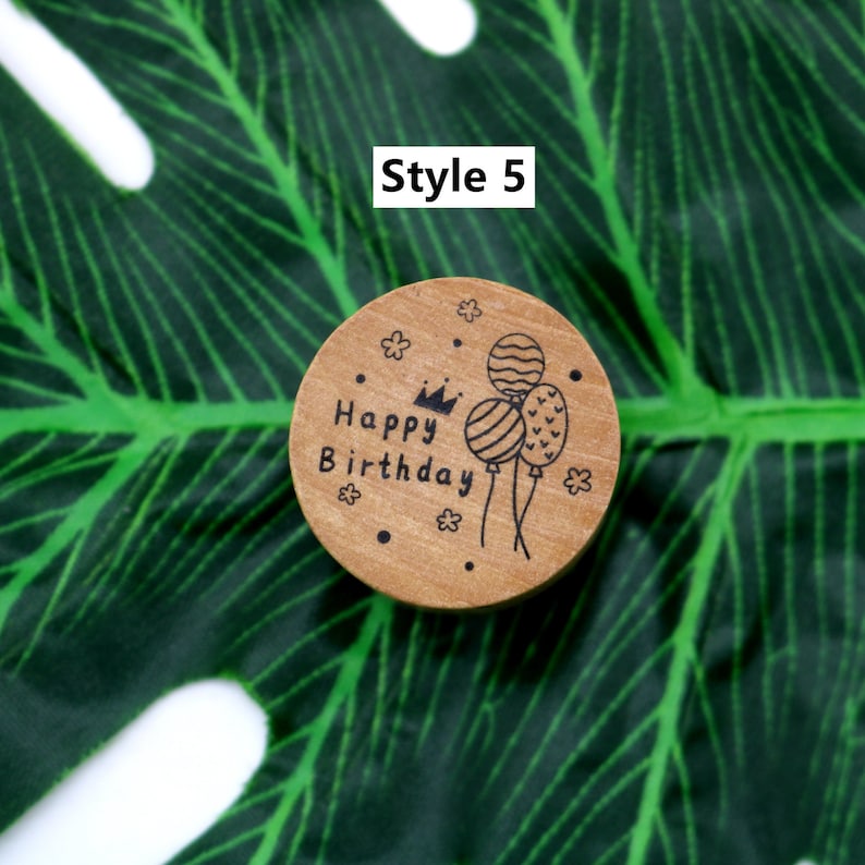 Puede incluir: Un sello de madera redondo con el texto "Happy Birthday" y una corona sobre la palabra "Happy". Tres globos est&aacute;n dibujados al lado del texto. El sello est&aacute; sobre un fondo de hoja verde.