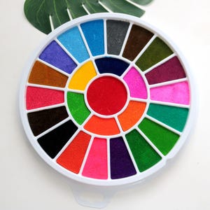 Könnte beinhalten: Ein runder, weißer Plastikbehälter mit 24 einzelnen Stempelkissen in verschiedenen Farben. Die Stempelkissen sind in einem Kreis angeordnet, wobei das mittlere Kissen rot ist. Der Behälter hat einen Griff an der Unterseite.