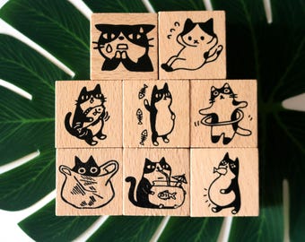May include: Nine wooden stamps with black cat illustrations. Each stamp features a different cat pose or expression, including cats with fish, in a bag, and using a hula hoop. The stamps are arranged on a green leaf.