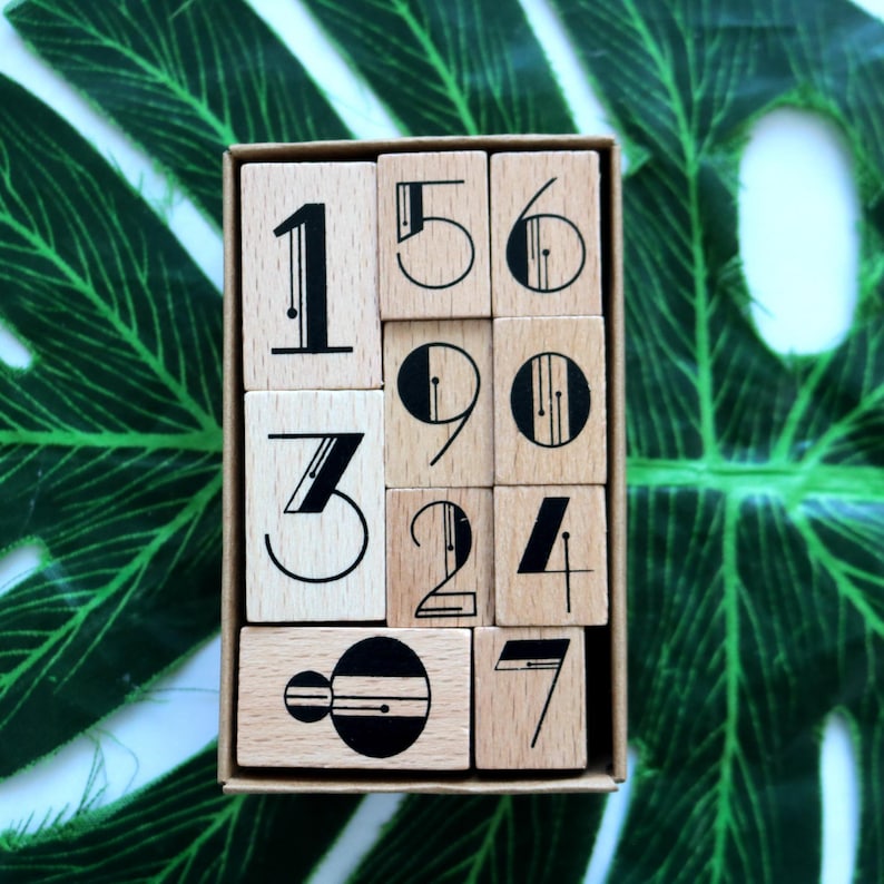 Puede incluir: Un juego de diez sellos de n&uacute;meros de madera en una caja. Los n&uacute;meros son 1, 2, 3, 4, 5, 6, 7, 8, 9 y 0. Cada n&uacute;mero est&aacute; estilizado con un dise&ntilde;o en blanco y negro.