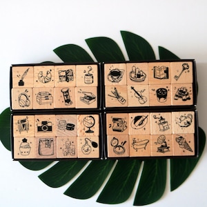 May include: A set of wooden stamps with black ink illustrations of various objects, including a croissant, headphones, a camera, a globe, a perfume bottle, and a bathtub. The stamps are arranged in black boxes, with a green leaf in the background.
