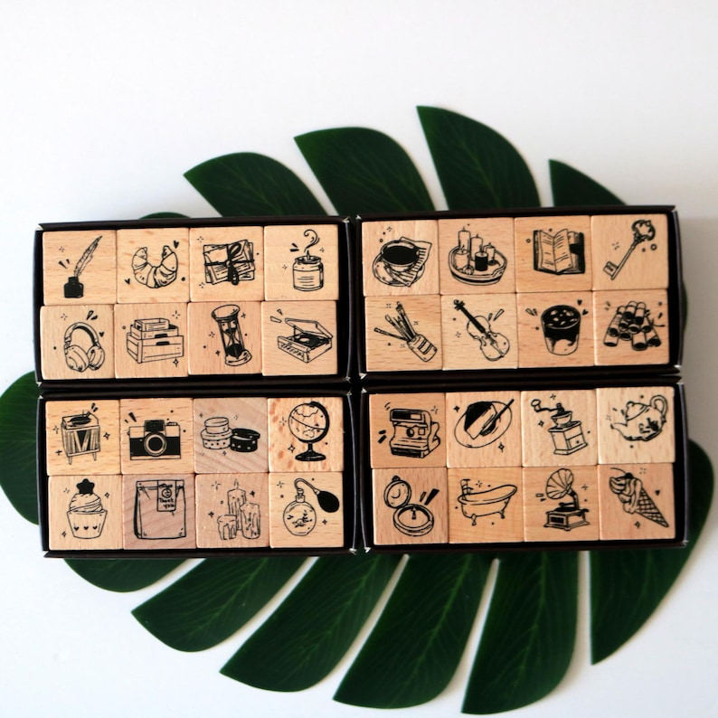May include: A set of wooden stamps with black ink illustrations of various objects, including a croissant, headphones, a camera, a globe, a perfume bottle, and a bathtub. The stamps are arranged in black boxes, with a green leaf in the background.