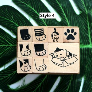 May include: A set of nine wooden stamps featuring various cat designs. The stamps are arranged in a 3x3 grid. The designs include a cat paw print, a cat with its tail up, a cat in a box, and a cat with its back to the viewer. The text "Style 4" is printed on the top of the image.