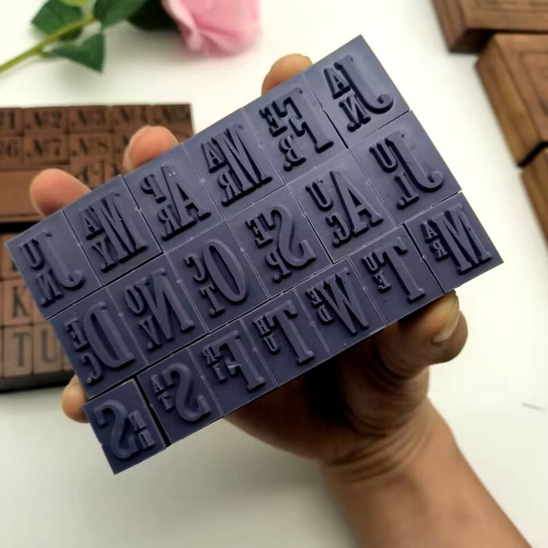 Puede incluir: Un juego de sellos de alfabeto de goma p&uacute;rpura con letras y n&uacute;meros. Los sellos est&aacute;n dispuestos en forma rectangular y se sostienen en una mano.