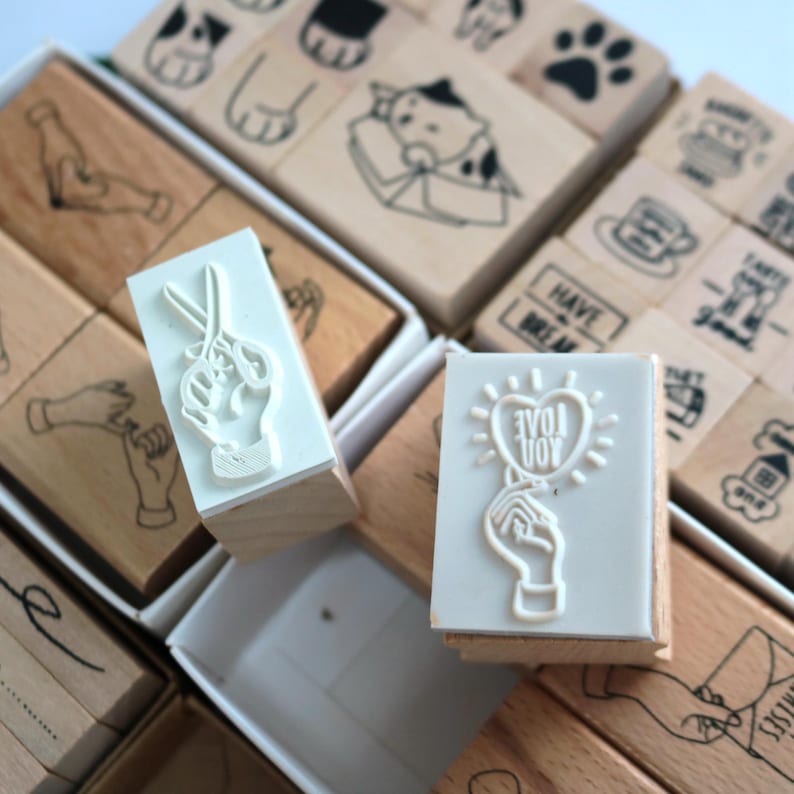 May include: A set of wooden stamps with various designs, including a hand holding scissors, a hand holding a heart with the text "love you", and a hand making a peace sign. The stamps are arranged in a grid pattern on a white surface.
