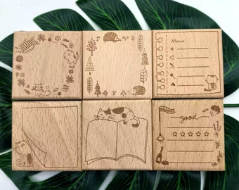 May include: Set of six wooden stamps with various designs, including a cat reading a book, a hedgehog in a forest, a cat in a wreath, a blank page, a memo pad, and a rating scale with the word "good".