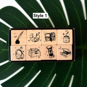 May include: A set of eight wooden stamps with black ink illustrations. The stamps feature a quill and inkwell, croissant, stack of books, headphones, hourglass, coffee, and a record player. The text "Style 1" is in a white box.