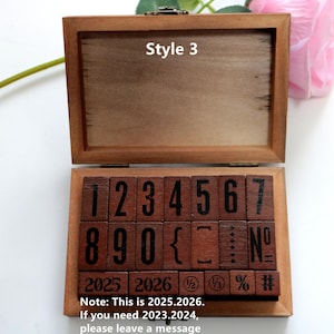 Puede incluir: Una caja de madera con tapa abatible, que contiene un juego de sellos de madera con n&uacute;meros, s&iacute;mbolos y los a&ntilde;os 2025 y 2026. La caja est&aacute; etiquetada como "Style 3".