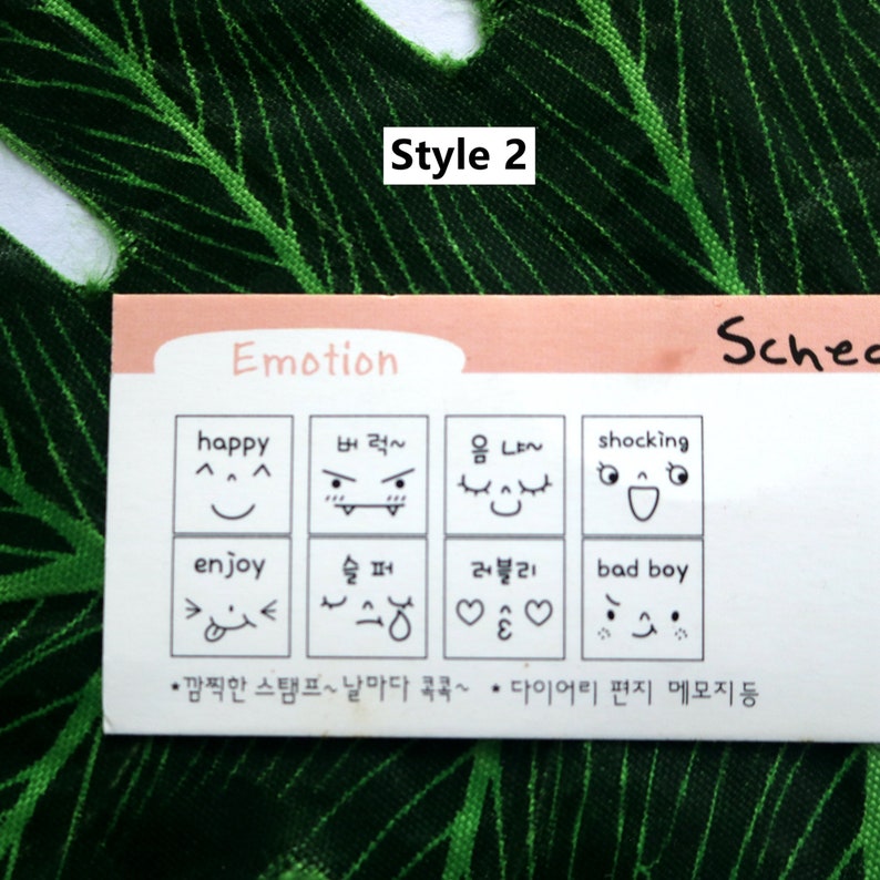 Op de afbeelding: Een witte kaart met de tekst "Style 2" in zwart bovenaan. De kaart heeft een roze achtergrond en bevat een raster van negen vierkanten. Elk vierkant bevat een ander cartoon gezicht met Koreaanse tekst eronder. De tekst op de kaart luidt "Emotion" en "Schec".