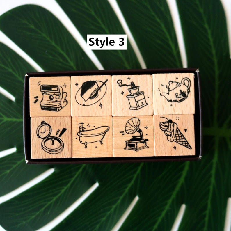 May include: A set of eight wooden stamps with black ink illustrations. The stamps feature various designs, including a camera, cake, coffee grinder, teapot, clock, bathtub, gramophone, and ice cream cone. The text "Style 3" is visible.
