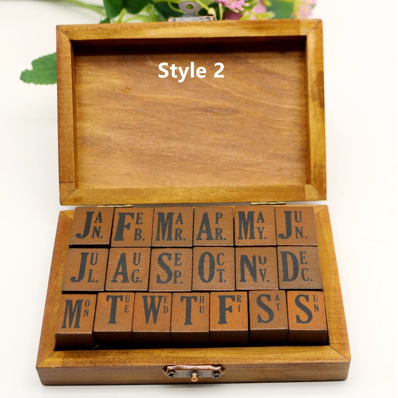 Puede incluir: Una caja de madera con tapa abatible que contiene un juego de sellos de letras y n&uacute;meros. Los sellos est&aacute;n dispuestos en filas y son de madera con tinta negra. La caja est&aacute; etiquetada como "Style 2".