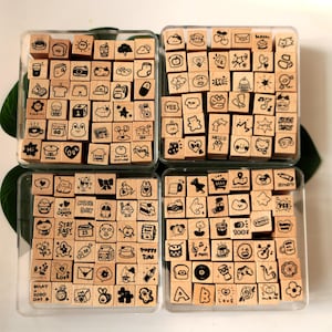 May include: A set of small, square wooden stamps featuring black ink illustrations of food, hearts, and text such as "Hello" and "Happy Time". The stamps are neatly arranged in clear plastic containers, perfect for crafting and scrapbooking projects.