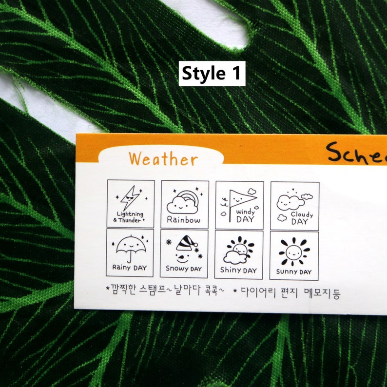 Op de afbeelding: Een witte kaart met het woord "Weather" in het zwart bovenaan gedrukt. De kaart is verdeeld in acht vierkanten, elk met een zwart-witte illustratie van een andere weersomstandigheid. De illustraties omvatten bliksem en donder, een regenboog, een winderige dag, een bewolkte dag, een regenachtige dag, een besneeuwde dag, een zonnige dag en een zonnige dag. De tekst "*깜찍한 스탬프~날마다 콕콕~ 다이어리 편지 메모지 등" staat onderaan de kaart gedrukt.