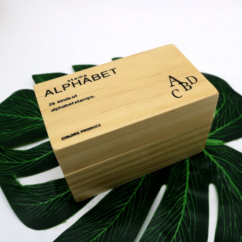 May include: A wooden box with a set of 26 alphabet stamps. The box is labeled "stamp alphabet 26 kinds of alphabet stamps. COLORS PRODUCE". The top of the box has the letters "A C B D" printed on it.