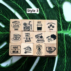 May include: A set of 12 wooden stamps with various designs, including a baguette, coffee cup, cake, and house. The stamps are arranged in a 4x3 grid on a green background. The text "Style 3" is printed above the stamps.
