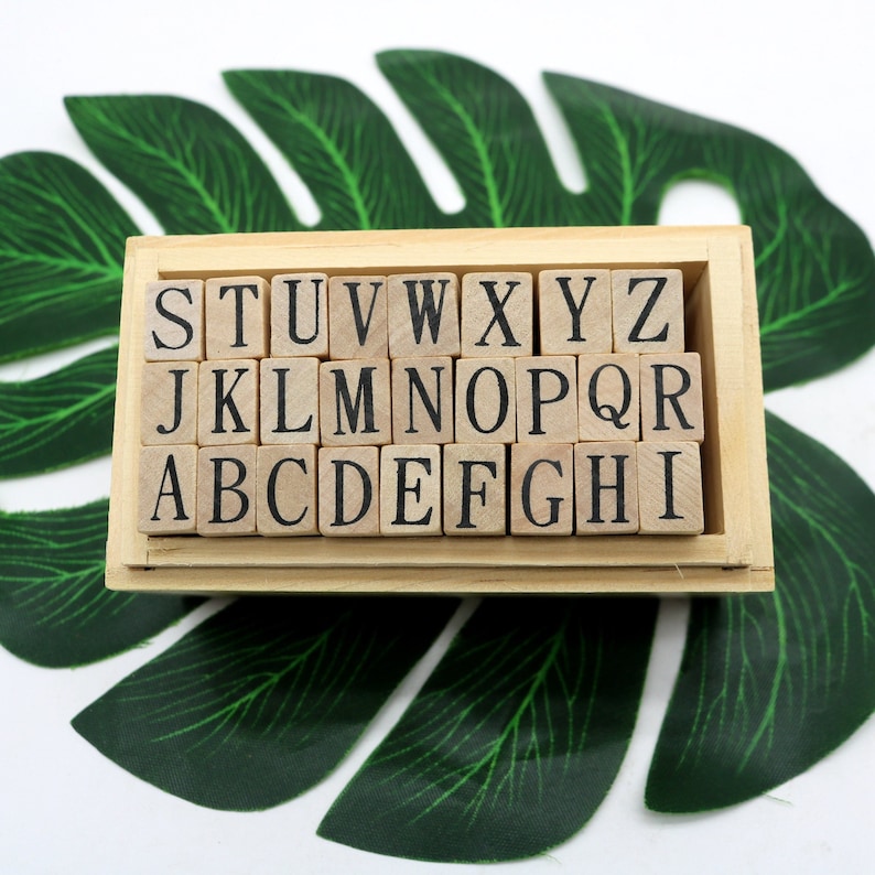 May include: A wooden box with a set of alphabet letter stamps. The stamps are arranged in alphabetical order, with the letters S, T, U, V, W, X, Y, and Z on the top row, J, K, L, M, N, O, P, and Q on the second row, and A, B, C, D, E, F, G, and H on the bottom row. The box is sitting on a green leaf.