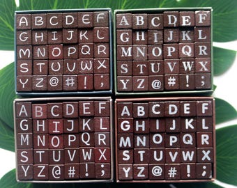 May include: Four sets of wooden alphabet stamps, each arranged in a grid within a small box. The letters are white, and the blocks are brown. Each set includes letters, numbers, and symbols.