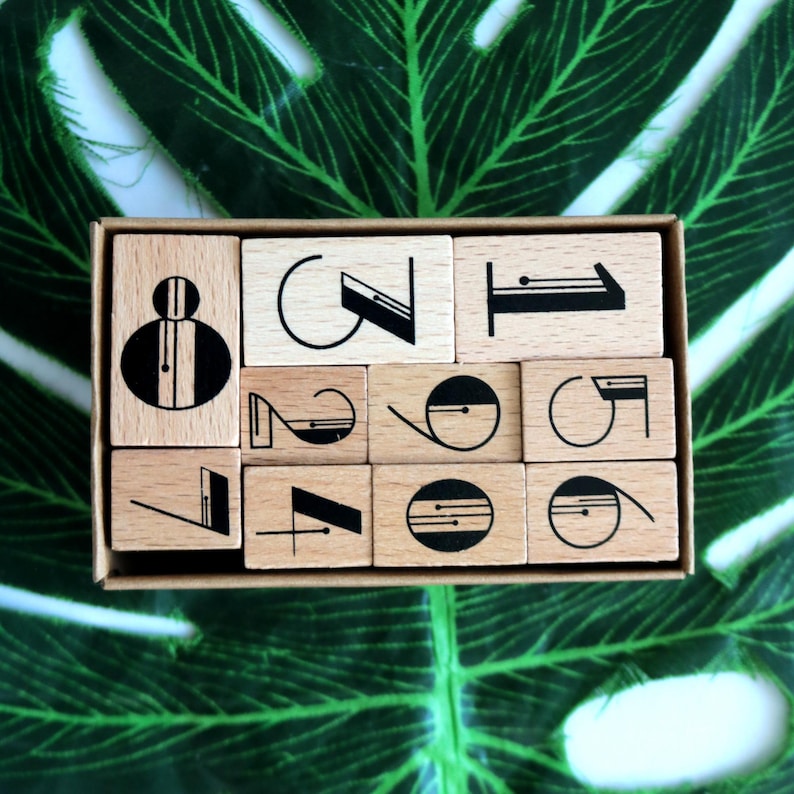 Puede incluir: Un juego de 12 sellos de alfabeto de madera con dise&ntilde;os geom&eacute;tricos en blanco y negro. Los sellos est&aacute;n dispuestos en una caja con fondo marr&oacute;n.