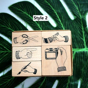 May include: Set of four wooden stamps featuring different hand gestures. The stamps depict a hand holding scissors, a hand holding a paintbrush, a hand pointing, and a hand holding a camera.