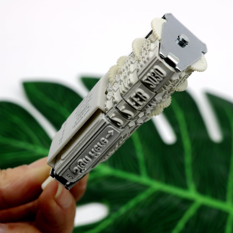 May include: A white plastic date stamp with a metal handle. The stamp has five wheels with numbers 0-9 and a sixth wheel with the text "20U H161-1".