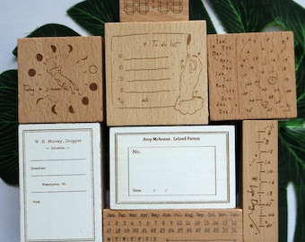 Pode incluir: Um conjunto de carimbos de madeira com vários desenhos, incluindo um carimbo de fase lunar, um carimbo de lista de tarefas, um carimbo de calendário, um carimbo de régua e um carimbo de etiqueta em branco. Os carimbos são todos feitos de madeira natural e têm um aspeto rústico.