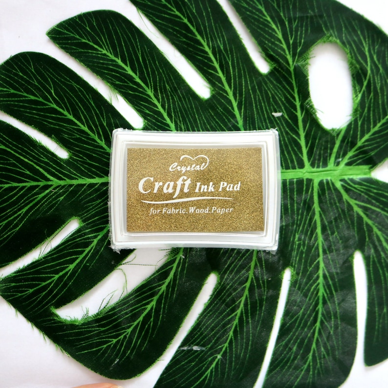May include: Gold craft ink pad with the text "Crystal Craft Ink Pad for Fabric. Wood. Paper" on a green leaf background.