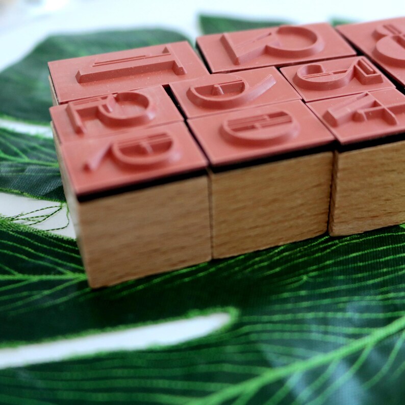 Puede incluir: Un conjunto de sellos de madera con letras de goma roja. Los sellos est&aacute;n dispuestos en forma rectangular y est&aacute;n sobre una hoja verde.