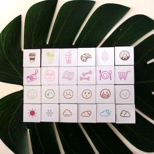 Peut inclure: Un ensemble de tampons carrés blancs avec diverses icônes, notamment une tasse de café, un croissant, une glace, un appareil photo, un masque facial, un livre, des haltères, une assiette et des couverts, un chariot de courses et des symboles météorologiques. Les tampons sont disposés sur une grande feuille verte.