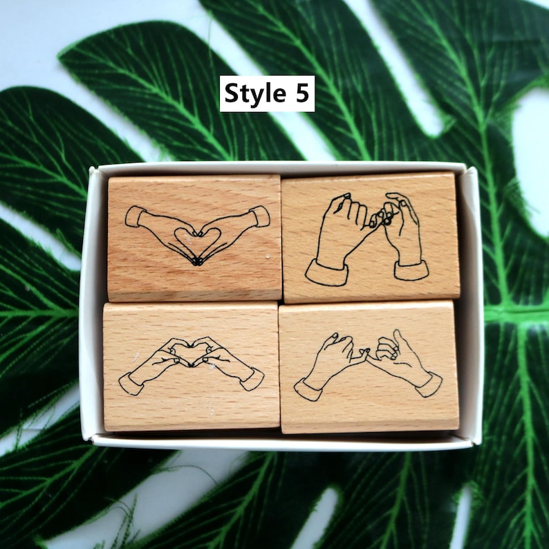 May include: Set of four wooden stamps with different hand gestures. The stamps are in a white box and feature designs of hands making a heart shape and pinky promises. The text "Style 5" is printed on the box.